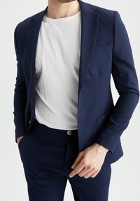 Marinblå strukturerad kavaj med slagkrage, öppen front och bröstficka, kombinerad med en vit T-shirt och matchande marinblå byxor.