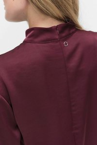 Bordowa bluzka z satyny z wysokim kołnierzem, zapinana z tyłu na kluczyk z ozdobnym srebrnym pierścieniem.