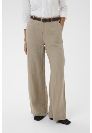 Persona lleva pantalones beige de pierna ancha con cinturón marrón, camisa blanca abotonada y zapatos negros de punta puntiaguda, de pie con una mano en el bolsillo.