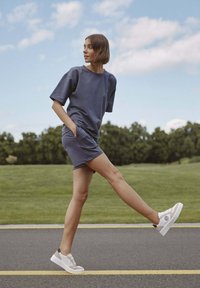 Tmavě modré šaty s krátkým rukávem a bočními kapsami, vyrobené z měkké látky. Spárované s bílými platformovými teniskami s černým akcentem, na zelené trávě.