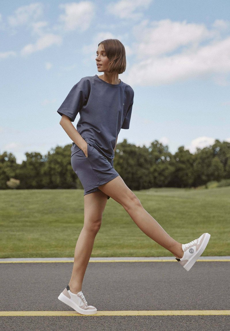 Tmavě modré šaty s krátkým rukávem a bočními kapsami, vyrobené z měkké látky. Spárované s bílými platformovými teniskami s černým akcentem, na zelené trávě.