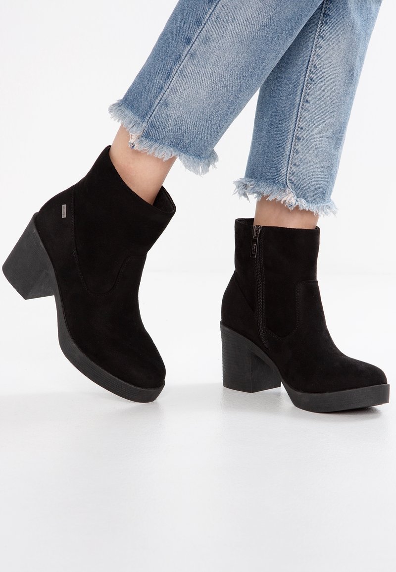 Bottines en daim noir à talon épais, avec une fermeture éclair latérale et une texture lisse. Portées avec un jean clair à ourlet effiloché.