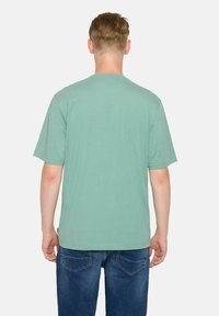 Tricou scurt din bumbac verde, cu guler rotund, croială lejeră. Textură ușoară, brand minimal la tiv. Asortat cu blugi denim de culoare bleumarin.