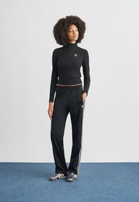 Conjunto deportivo negro que incluye una parte superior ajustada de manga larga con cremallera y pantalones de pierna ancha con tres franjas blancas y detalles de logotipo.