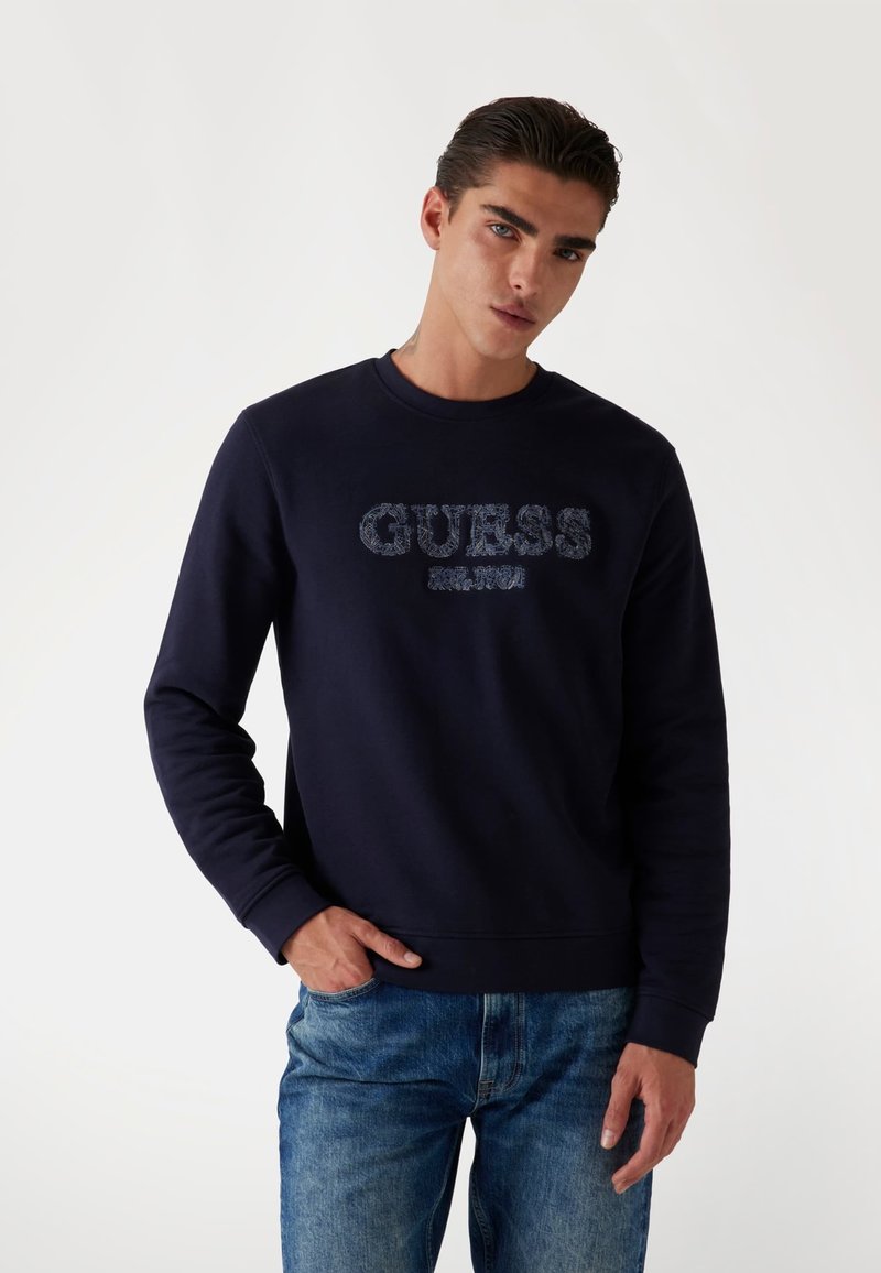 Felpa blu navy con collo a giro e maniche lunghe. La parte frontale presenta un logo "GUESS" testurizzato in un colore a contrasto.