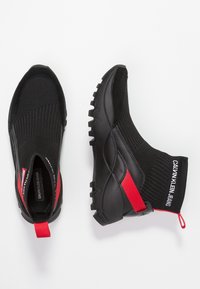 Černé tenisky na slip-on s červenými poutky na patě, texturovaný pletený svršek, silné robustní podrážky a značka "CALVIN KLEIN JEANS" umístěná na límci kolem kotníku.