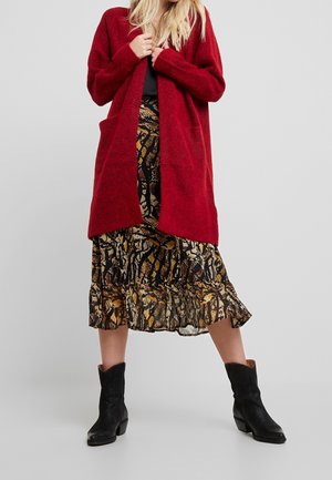 Vrouw draagt een lange rode cardigan, een midi-rok met slangenprint, een zwart topje en zwarte enkelboots tegen een effen grijze achtergrond.