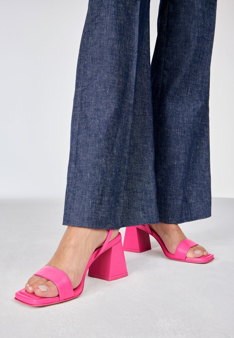 Sandali con tacco alto rosa, con punte quadrate, cinturini larghi e un tacco geometrico spesso, abbinati a pantaloni a gamba larga in denim scuro.
