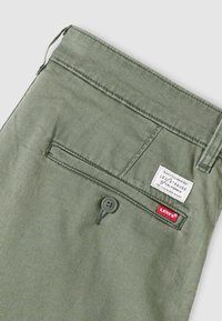 Πράσινη τσέπη παντελονιού Levi's με κούμπωμα κουμπιού, κόκκινη ετικέτα Levi's και λευκή ετικέτα με το λογότυπο ραμμένη πάνω από την τσέπη.