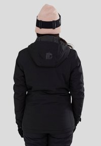 Persoon gekleed in een zwarte winterjas met capuchon en zwarte handschoenen, met de rug naar de kijker, en een lichtroze gebreide muts met een zwarte band.