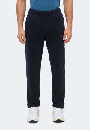 Homme portant un pantalon de survêtement bleu marine, un t-shirt bleu et des chaussures de sport blanches, debout les bras détendus le long du corps.