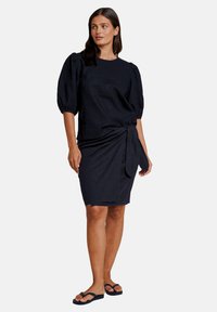 Mørkeblå kjole med pufferm, rund utringning og knyttedetalj i midjen. Laget av et teksturert stoff.