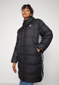 Juodas pūkinis paltas su užtrauktuku priekyje, aukštu apykakle, šoninėmis kišenėmis ir baltais dryžiais ant rankovių. Ypatumai: siuvinėti raštai ir Adidas logotipas.