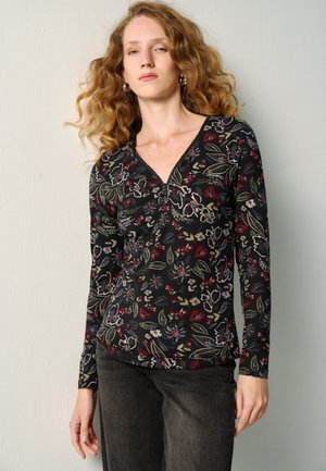 Mujer con cabello rizado rojo que lleva una blusa negra de manga larga con flores, con cremallera en el escote y pantalones oscuros, de pie contra una pared clara.