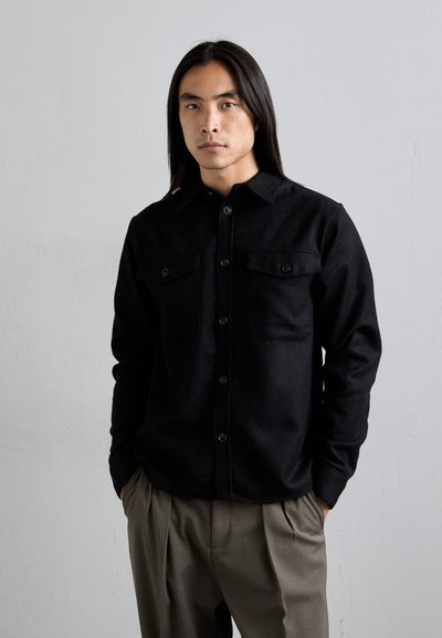 Chemise noire à boutons avec col, manches longues et deux poches poitrine. Fabriquée en tissu texturé avec une coupe ajustée.