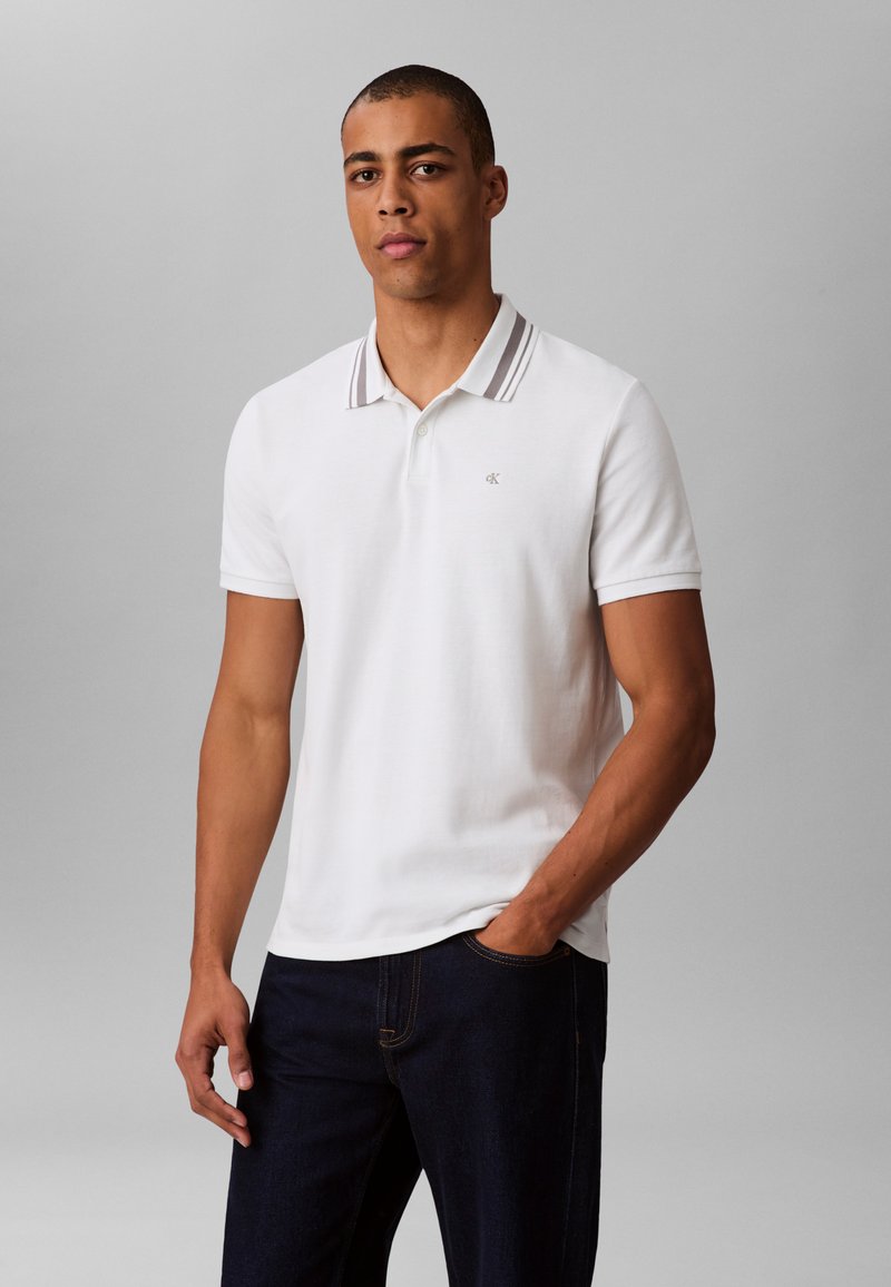 White polo shirt with a collar featuring gray stripes, short sleeves, and a small logo on the chest; paired with dark jeans.