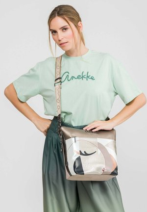 Frau mit hellgrünem "Anekke" T-Shirt und dunkelgrüner Hose, die eine metallische Umhängetasche mit abstraktem Gesichtsmotiv hält.