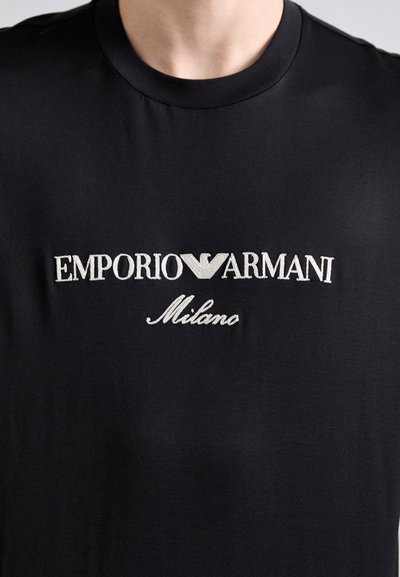 T-shirt en coton noir avec logo brodé blanc "EMPORIO ARMANI Milano" en police serif, col rond et texture lisse.