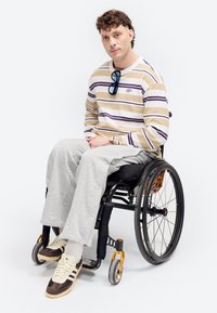 Άνδρας με σγουρά μαλλιά που φοράει μία ριγέ μπλούζα με μακριά μανίκια και γκρι παντελόνι, καθισμένος σε μια μαύρη αναπηρική καρέκλα με λευκό φόντο.