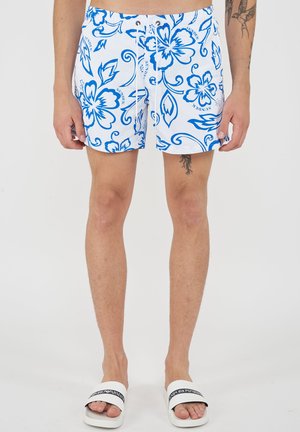 Man draagt witte zwemshorts met blauwe tropische bloemenprint en witte Emporio Armani slippers, toont tattoo op linker dij en rechter arm.