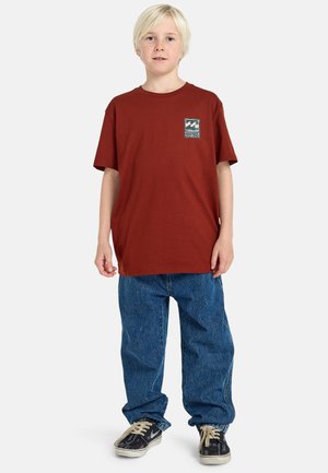 T-shirt en coton bordeaux avec un patch graphique rectangulaire sur la poitrine gauche ; associé à un jean en denim bleu ample et des baskets noires.