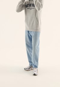 Hombre con jeans azul claro, sudadera gris con capucha "Newport" y zapatillas blancas de correr, de pie sobre un fondo claro y simple.