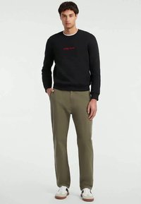 Mladý muž stojí oblečený v černé mikině s červeným logem "Guess Jeans", olivově zelených kalhotách a bílých teniskách.