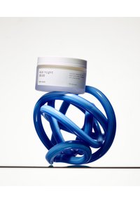 Hvit krukke med skrulokk merket "mid/natt kroppsbalsam," plassert oppå en vridd blå plasttråd, som reflekterer en glatt tekstur.