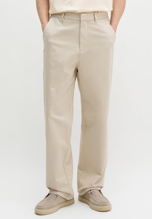 Sirged beežid püksid koos beežide seemisnahksete kingadega, käed taskutes, seistes helel lihtsal taustal.