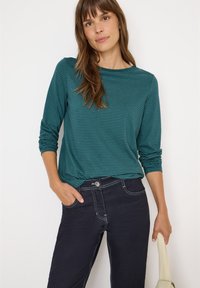 Camicia a maniche lunghe a righe turchesi con scollo rotondo, abbinata a jeans scuri con cuciture bianche e una finitura liscia. Look casual.