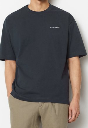 Person wearing a plain black Marc O'Polo t-shirt and beige pants, standing with one hand in pocket against a light background.