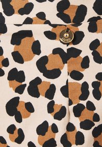 Lähi-kuva vaaleasta kankaasta, jossa on musta ja ruskea leopardikuosi sekä pieni pyöreä kultaketju, jossa on kristalli, ommellussa saumassa.