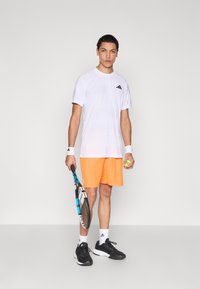 Camiseta deportiva blanca con textura y logo negro, pantalones cortos naranjas, zapatos negros, muñequeras, raqueta de tenis y pelota de tenis amarilla.