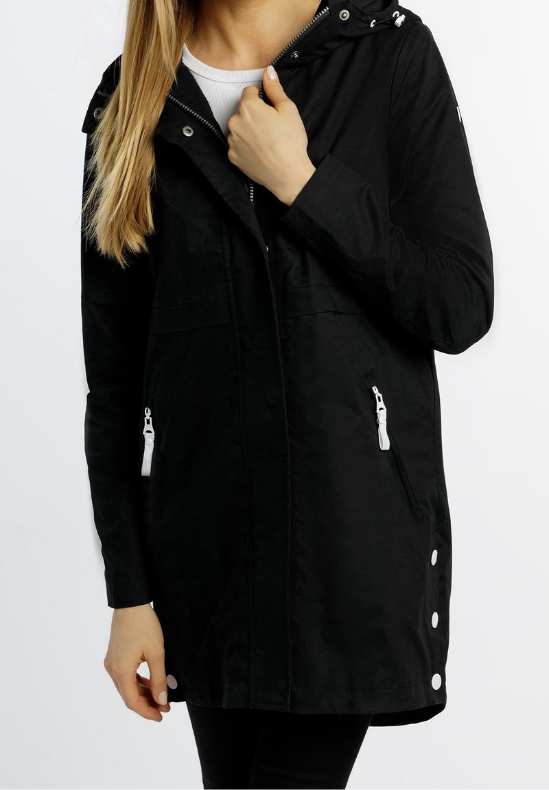 Femme portant une veste à capuche noire avec des tirettes de fermeture éclair blanches, tenant le col de la veste, sur un fond blanc uni.