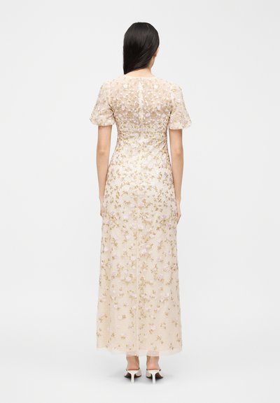 Γυναίκα με μακριά σκούρα μαλλιά, που φοράει μακρύ μπεζ φόρεμα με κεντήματα λουλουδιών και κοντά φουσκωτά μανίκια, στέκεται γυρισμένη προς τα πίσω.