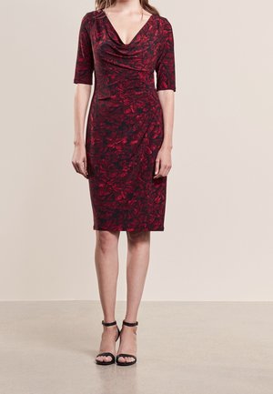Frau trägt ein figurbetontes, knielanges, bordeauxrotes Kleid mit Blumenmuster, Ellbogen langen Ärmeln, Wasserfallausschnitt und schwarzen Pumps mit Knöchelriemen.