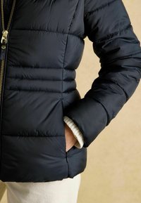 Osoba oblečená v čiernej prešívanej bunde so zlatým zipsom, s jednou rukou v bočnej vrecku a krémovými nohavicami na béžovom pozadí.