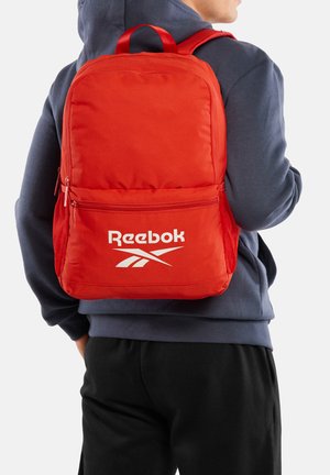 Osoba nosząca jasnoczerwoną torbę Reebok z zapinanymi na zamek kieszeniami, siateczkowymi bocznymi kieszeniami i regulowanymi paskami na ramiona, ubraną na ciemną bluzę z kapturem.