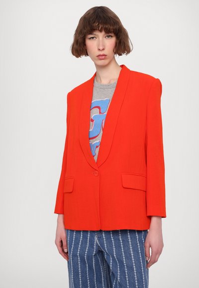 Bright red blazer with a slight sheen, notched lapels, single button closure, and two front pockets. Worn over a gray graphic t-shirt.