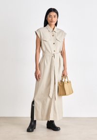 Μπεζ, αμάνικο midi φόρεμα με κολάρο και ζώνη. Διαθέτει δύο τσέπες στο στήθος, πλαϊνές σχισμές και ένα μοντέλο που κρατάει μία μικρή κίτρινη τσάντα.