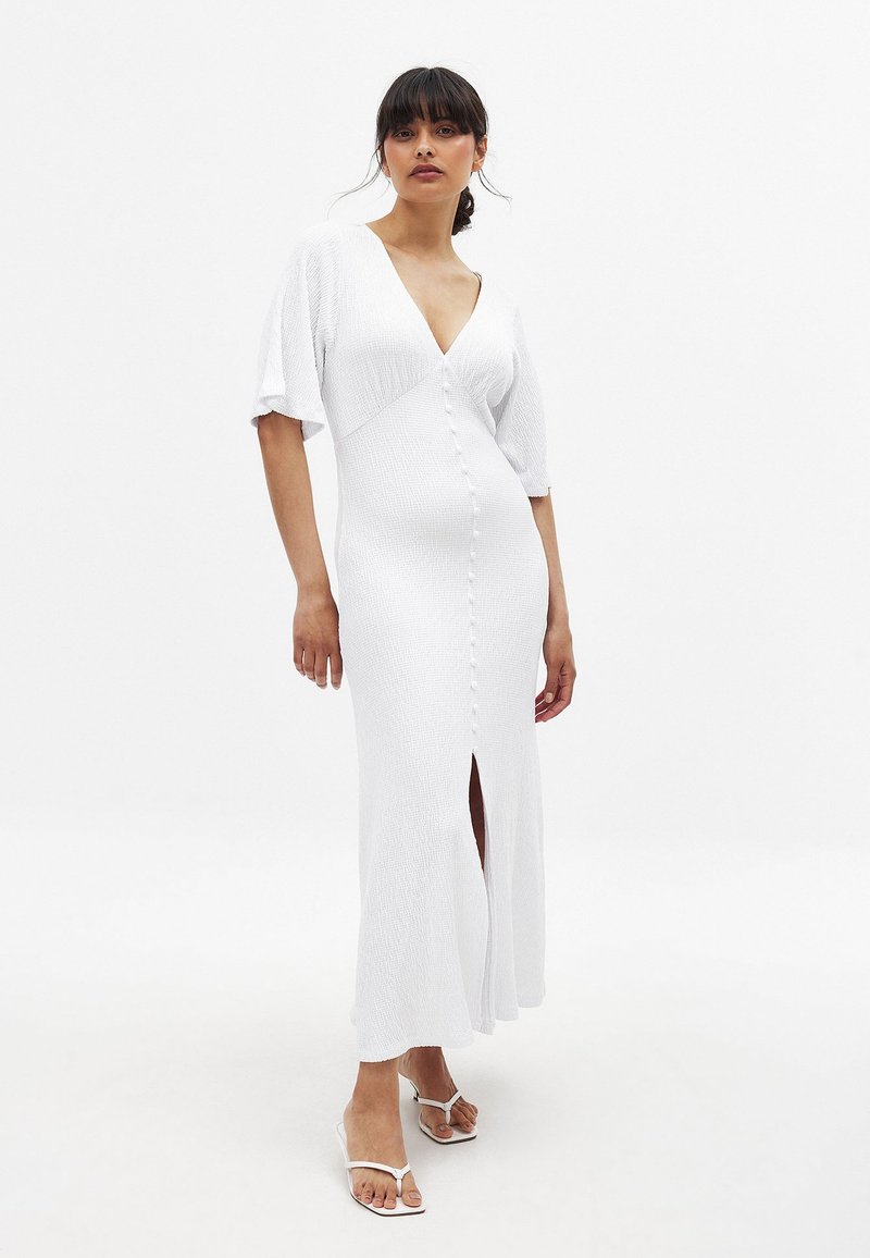 Robe blanche, longueur genoux, avec un décolleté en V, des manches courtes et un tissu texturé. Présente des boutons sur le devant et une fente sur le côté. Portée avec des sandales blanches.