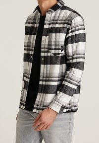 Homme portant une chemise à carreaux noire et blanche boutonnée par-dessus un haut noir et un pantalon gris clair, debout devant un fond uni.