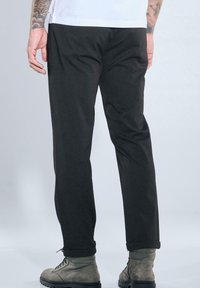 Mann trägt schwarze Hose, graue Schnürstiefel und weißes Hemd, von hinten gesehen, mit sichtbaren tätowierten Armen.