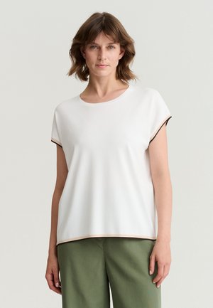 Vrouw met kort bruin haar, gekleed in een wit T-shirt met korte mouwen met lichtbruine afwerking en olijfgroene broek, staand tegen een effen achtergrond.