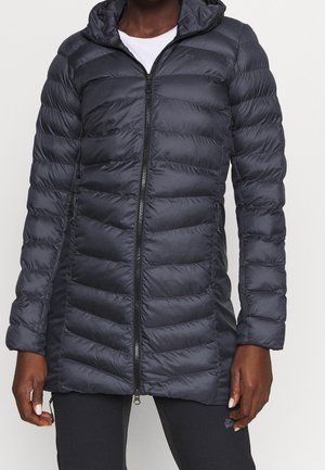 Zwarte gewatteerde hooded jas met ritssluiting aan de voorkant, gedragen over zwarte broek, zichtbaar van nek tot midden dij op een persoon met een donkere huidtint.