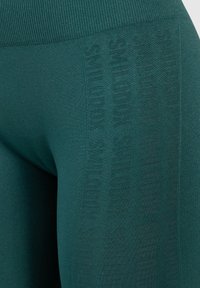Turkusowe legginsy z żebrowanym pasem i subtelnym wytłaczanym logo. Materiał wydaje się elastyczny o gładkiej fakturze.