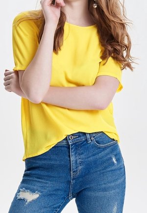 Donna con capelli lunghi che indossa una maglietta gialla a maniche corte e jeans blu strappati, con le braccia incrociate, in piedi contro uno sfondo chiaro e uniforme.