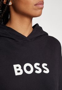 Gros plan d'une personne portant un sweat à capuche noir avec le mot « BOSS » en lettres blanches en gras sur la poitrine et de petites boucles d'oreilles rondes argentées.