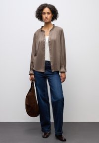 Hnědá košile na knoflíky s volánovým límcem, kombinovaná s modrými širokými džíny a hnědými koženými botami, držící hnědou tašku.