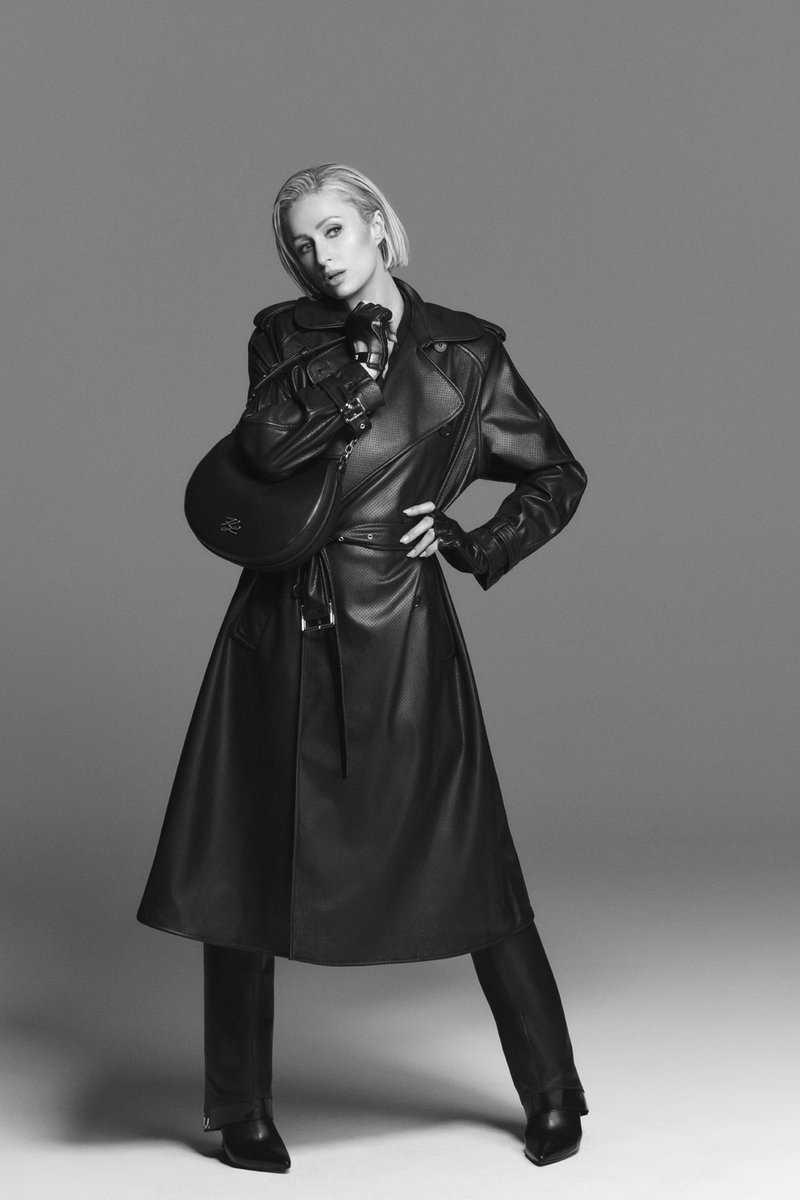 Μαύρο δερμάτινο παλτό τρεντς με υφή, εφαρμοστά μανίκια και ζώνη που σφίγγει στη μέση· ταιριαστά μποτάκια με ψηλό τακούνι και στρογγυλή τσάντα χειρός.